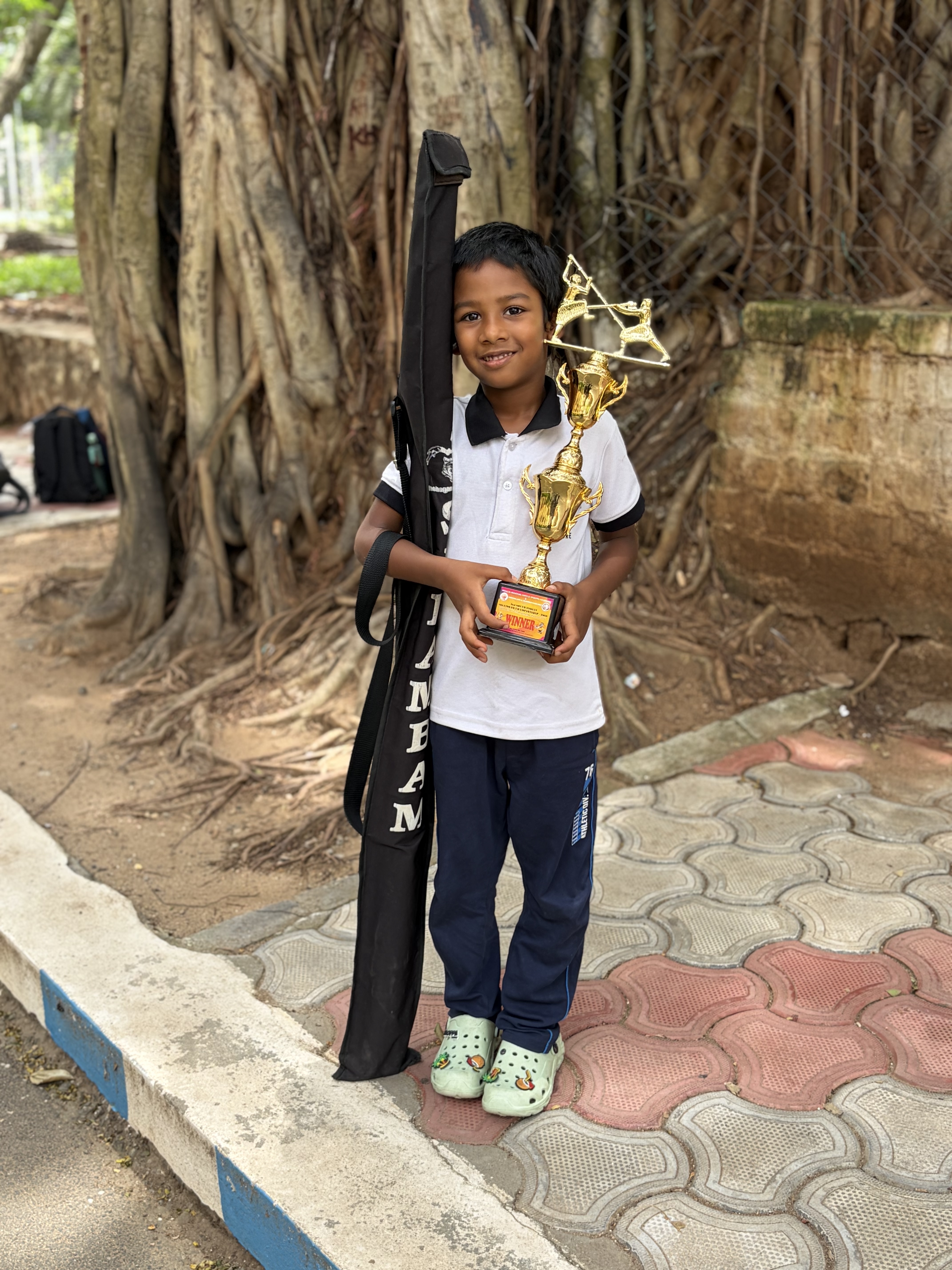 Prithvi standing proud under a banyan tree with his trophy and Silambam stick bag
